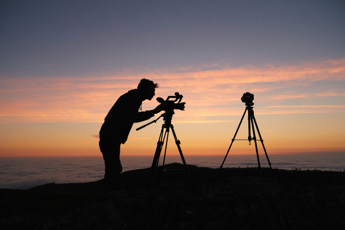 Silhouette of a videographer adjusting a camera on a tripod at sunset, with a second camera on a tripod beside him, above a sea of clouds